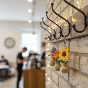 May include: A decorative wall-mounted coat rack with five dark metal hooks. Three crochet flower keychains hang from the hooks. The wall is made of light-colored bricks, and small lights are strung along the wooden backing.