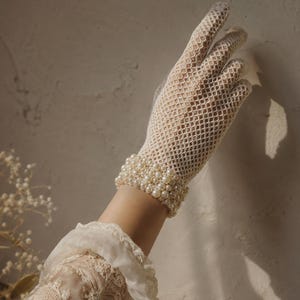 May include: A cream-coloured fishnet glove featuring a wrist cuff adorned with multiple rows of small, round, white faux pearls. The glove has a diamond-shaped mesh pattern and covers the hand and fingers, extending to the wrist area.