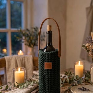 Pode incluir: Um porta-garrafas de malha verde-escura com uma alça de couro castanho e um emblema retangular de couro com o texto em relevo Willow & Fern. O suporte contém uma garrafa de vinho de vidro com rolha de cortiça, proporcionando um transporte seguro.