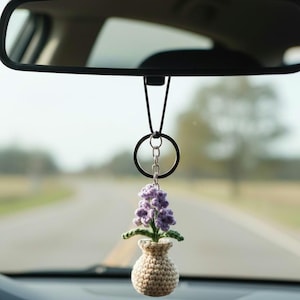 May include: A crocheted car charm featuring a small vase with a purple flower arrangement. The vase is beige, and the flower has green leaves. The charm hangs from a black ring and chain, suspended from a car's rearview mirror.