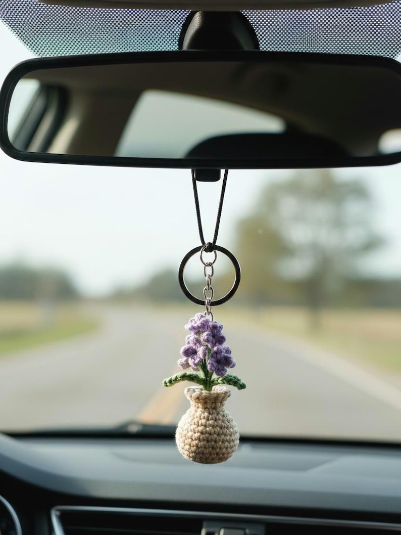 May include: A crocheted car charm featuring a small vase with a purple flower arrangement. The vase is beige, and the flower has green leaves. The charm hangs from a black ring and chain, suspended from a car's rearview mirror.
