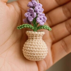 May include: A handmade crochet keychain featuring a miniature flower arrangement. The keychain showcases a beige vase holding a cluster of purple flowers with green leaves. The keychain is attached to a silver ring.
