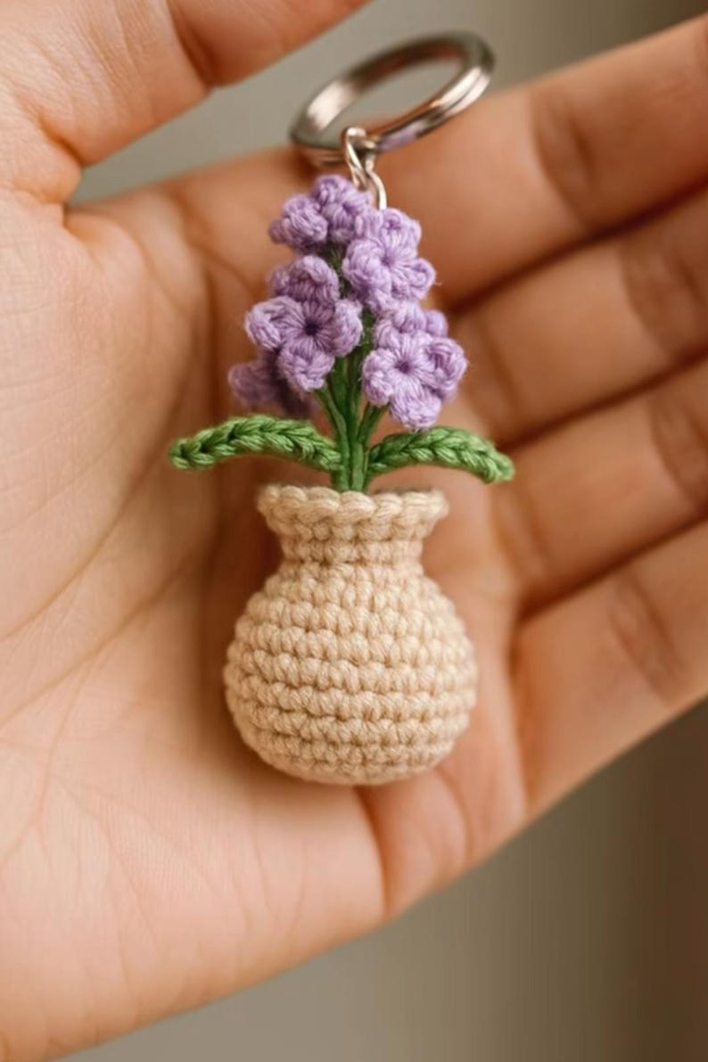 May include: A handmade crochet keychain featuring a miniature flower arrangement. The keychain showcases a beige vase holding a cluster of purple flowers with green leaves. The keychain is attached to a silver ring.