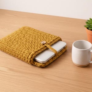 May include: Mustard yellow crocheted laptop sleeve with a wooden button closure. A silver laptop is partially visible inside the sleeve. A white ceramic mug and a small potted succulent are also on the wooden surface.