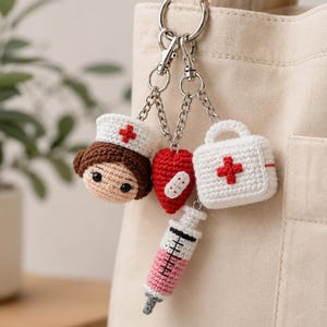 Peut inclure: Un porte-clés en crochet composé d'une figurine d'infirmier aux cheveux bruns avec une coiffe blanche à croix rouge, un cœur rouge avec un pansement, une trousse médicale blanche à croix rouge et une seringue à liquide rose.