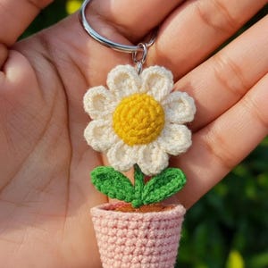 May include: Crocheted flower keychain featuring a white daisy with a yellow center, green leaves, and a pink pot. The keychain is attached to a silver ring. The handmade item is held in a hand.