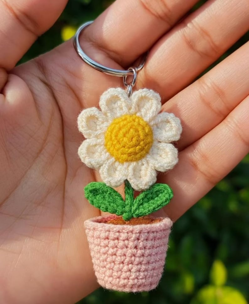 May include: Crocheted flower keychain featuring a white daisy with a yellow center, green leaves, and a pink pot. The keychain is attached to a silver ring. The handmade item is held in a hand.