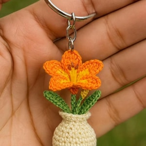 May include: A handmade crochet keychain featuring a vibrant orange flower with yellow accents, green leaves, and a small beige vase. The flower is attached to a silver keyring, creating a charming and unique accessory.