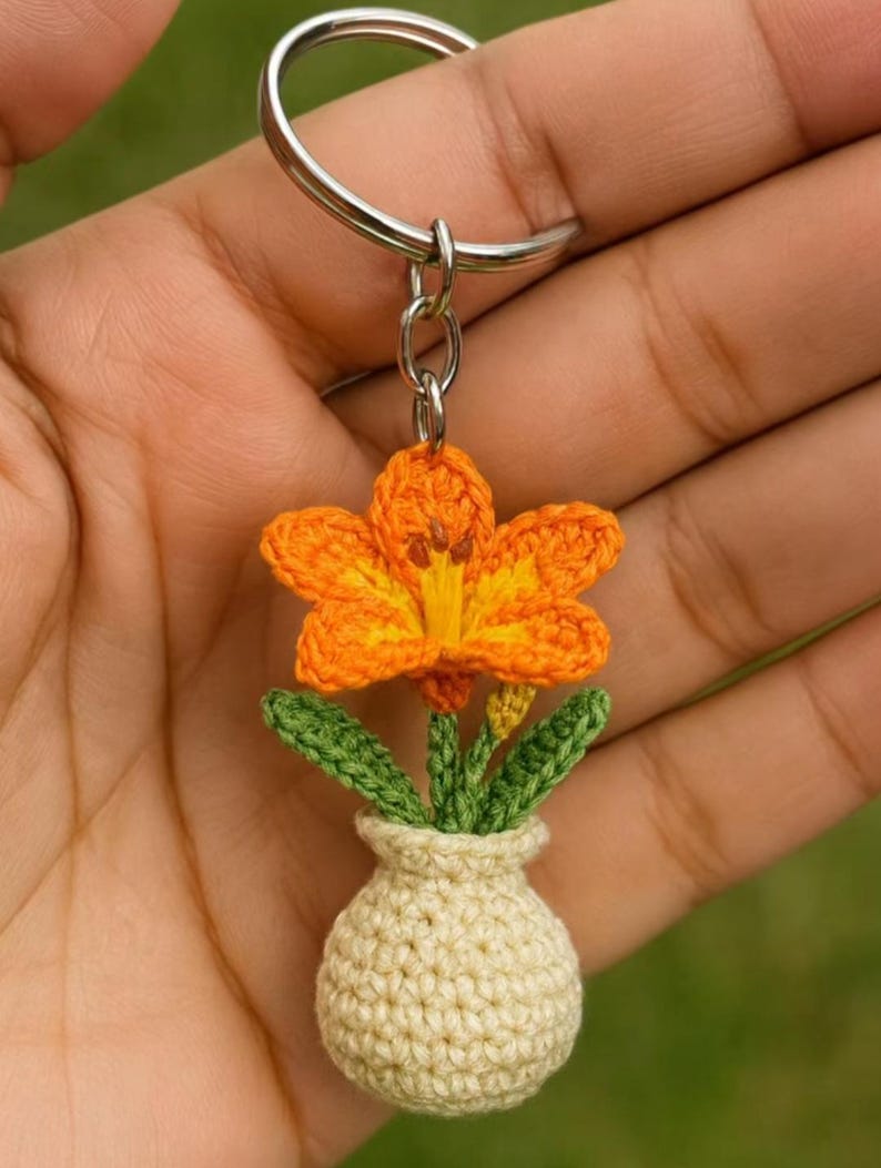 May include: A handmade crochet keychain featuring a vibrant orange flower with yellow accents, green leaves, and a small beige vase. The flower is attached to a silver keyring, creating a charming and unique accessory.