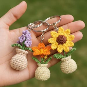 May include: Three crochet flower keychains: a purple hyacinth, an orange lily, and a yellow sunflower. Each flower is in a beige vase with green leaves. The keychains are held in a hand against a blurred green background.