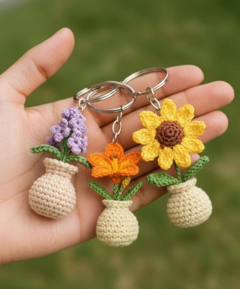 May include: Three crochet flower keychains: a purple hyacinth, an orange lily, and a yellow sunflower. Each flower is in a beige vase with green leaves. The keychains are held in a hand against a blurred green background.