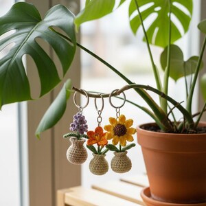 May include: Three crochet flower keychains: a purple hyacinth, an orange flower, and a yellow sunflower. Each flower is in a small beige vase. The keychains hang from a plant branch, with a potted plant in the background.