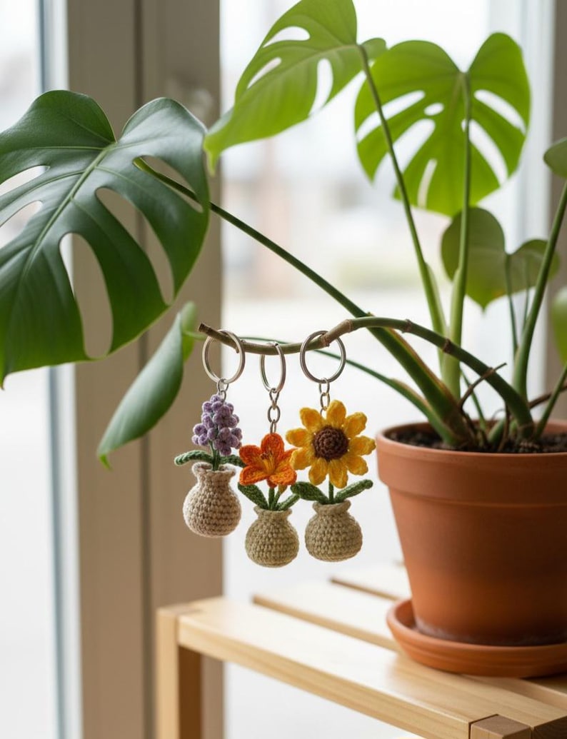May include: Three crochet flower keychains: a purple hyacinth, an orange flower, and a yellow sunflower. Each flower is in a small beige vase. The keychains hang from a plant branch, with a potted plant in the background.