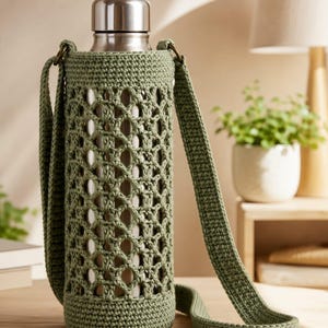 May include: A stainless steel water bottle encased in a sage green crocheted sleeve with an openwork lattice pattern and a matching long shoulder strap. The bottle features a smooth metallic finish and a rounded silver cap at the top.