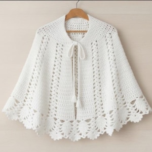 Può includere: Una mantella bianca all'uncinetto con orlo smerlato e chiusura con fiocco. La mantella è realizzata in un materiale morbido e a trama aperta ed è appesa a una gruccia di legno. La lunghezza della mantella è di circa 63,5 cm.