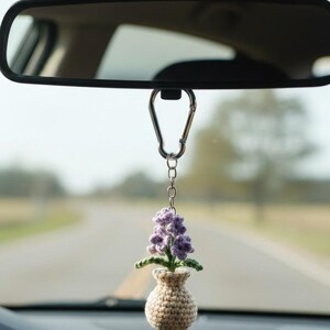 May include: A crocheted car charm featuring a small beige vase with a purple flower arrangement. The charm hangs from a silver carabiner attached to the rearview mirror inside a car, with a blurred road in the background.