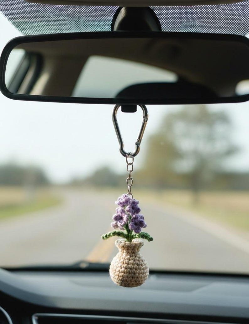 May include: A crocheted car charm featuring a small beige vase with a purple flower arrangement. The charm hangs from a silver carabiner attached to the rearview mirror inside a car, with a blurred road in the background.