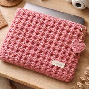 Könnte beinhalten: Eine rosa gehäkelte Laptoptasche mit strukturiertem Muster. Ein kleiner gehäkelter Herzanhänger hängt an einem goldfarbenen Schlüsselanhänger. Die Hülle hat ein kleines Etikett mit der Aufschrift "handmade".