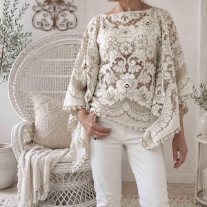 May include: A cream-colored lace top with a floral pattern. The top has a boat neck and wide sleeves. The model is wearing white pants. The background includes a white wicker chair, a throw blanket, and a potted plant.