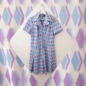 May include: A vintage dress with a diamond pattern in purple and blue. The dress has a collared neckline, a front zip, and two front pockets. White trim accents the collar, pockets, and cuffs. The dress is hanging on a white hanger. The background features a diamond pattern.