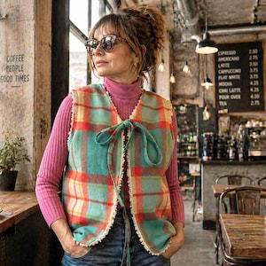 May include: A colorful plaid vest with a teal base and red, orange, and yellow accents. The vest has a tie closure and a cream-colored trim. The person is wearing a pink turtleneck and sunglasses. The image is taken in a coffee shop.