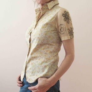 Può includere: Camicia abbottonata con motivo floreale giallo, rosa e blu. Le maniche corte sono trasparenti, con decorazioni floreali di perline. La camicia è abbinata a jeans blu.