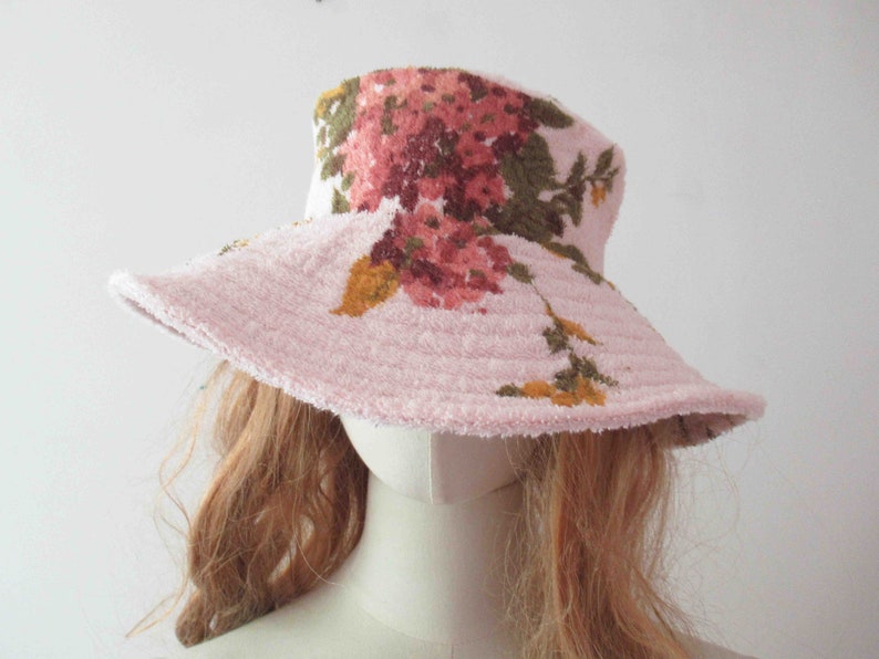 May include: A pink floral bucket hat with a wide brim. The hat is made of a soft, textured fabric and features a floral print in shades of pink, green, and yellow.