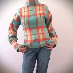 May include: A teal, orange, and yellow plaid sweater with a high neck. The sweater has long sleeves and a fitted shape. The person is wearing blue jeans.