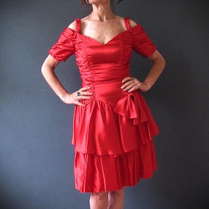May include: A vibrant red dress with a V-neckline and off-the-shoulder sleeves. The dress features a ruched bodice, a tiered skirt, and a bow detail at the waist. The dress is made of a shiny, satin-like material.