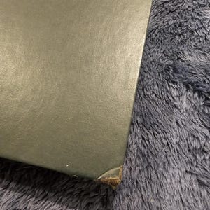 May include: A corner of a dark green, possibly leather-bound book or box. The edges are slightly worn, with a visible brown material at the corner. The background is a fuzzy, dark blue surface.