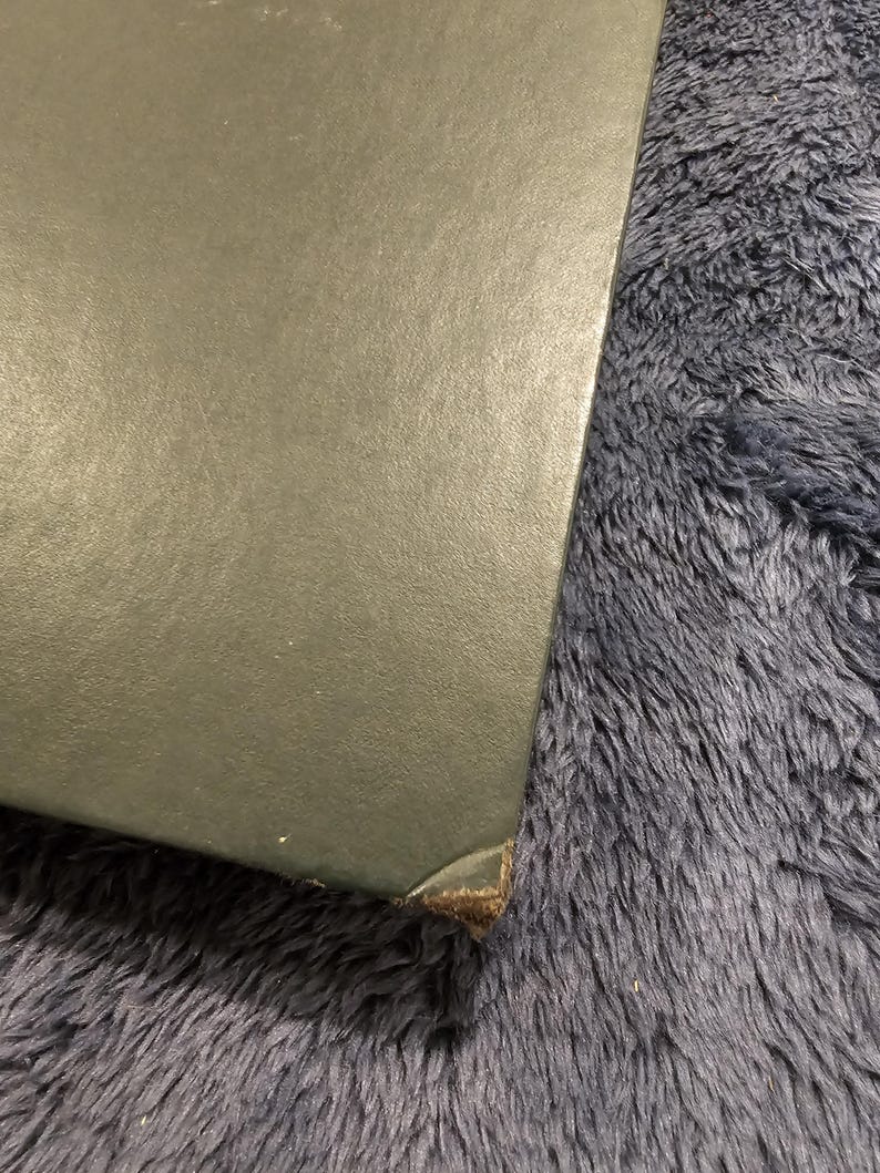 May include: A corner of a dark green, possibly leather-bound book or box. The edges are slightly worn, with a visible brown material at the corner. The background is a fuzzy, dark blue surface.