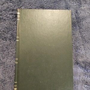 May include: A hardcover book with a dark green cover. The spine is visible with gold accents. The book is rectangular and appears to be in good condition. The background is a textured, dark blue surface.
