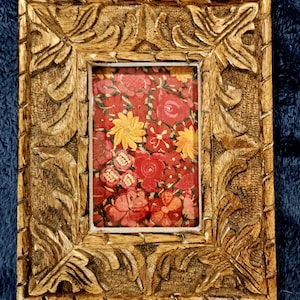 May include: A rectangular, wooden picture frame with intricate carved floral designs. The frame surrounds a floral print with red, yellow, and pink flowers on a red background. The frame has a natural wood finish.