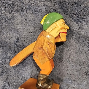 May include: Wooden nutcracker in the shape of a man, wearing a green and yellow hat, brown jacket, orange pants, and black boots. The nutcracker has a lever arm and is mounted on a wooden base. The figure has a red mouth.