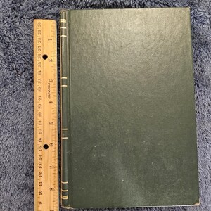 May include: A dark green book with a ruler alongside it. The book has a textured cover and a spine with vertical lines. The ruler is marked in inches, showing measurements up to 30 inches.