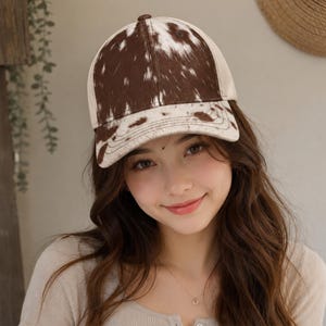 May include: A baseball cap featuring a brown and white cow print pattern across the front panels and the brim. The cap has a structured crown with a button on top and a curved visor, showcasing a distinct animal-inspired hide texture design.