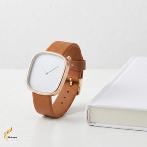 Könnte beinhalten: Eine minimalistische Armbanduhr mit quadratischem, goldfarbenem Zifferblatt und weißem Zifferblatt. Die Uhr hat ein braunes Lederarmband und eine goldene Schnalle. Die Uhr wird neben einem weißen Buch auf einer weißen Oberfläche präsentiert.