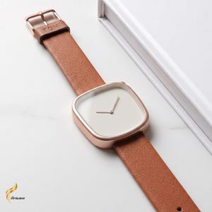 May include: A minimalist wristwatch with a square face and a brown leather strap. The watch has a rose gold case and a white dial with simple hands. The strap is secured with a rose gold buckle. The word "TOMI" is engraved on the buckle.