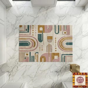 May include: A rectangular rug with a geometric design in beige, pink, teal, and gold. The rug features arched and rectangular shapes. The text "Avika Rugs" is visible on the rug. The rug is placed on a white marble surface.