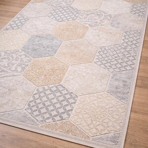 Peut inclure: Tapis rectangulaire à motifs géométriques. Il présente une série d'hexagones beiges, gris et bronzés, chacun avec un motif décoratif différent, créant un aspect texturé. Le tapis est bordé d'une bordure crème.