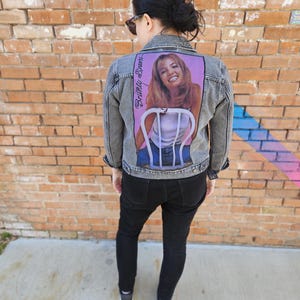 May include: A grey denim jacket with a faded appearance, showcasing a large graphic on the back. The graphic is a portrait of a woman seated on a chair, with the text "Britney Spears" in cursive. The jacket has a standard collar and button fastenings.