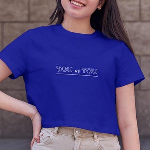 May include: A royal blue crop top with the text "YOU vs. YOU" in white. The shirt is paired with beige wide-leg pants. The image is taken outdoors, with a stone building and steps in the background.