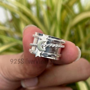 May include: A silver spinner ring featuring a crescent moon and star design. The ring has a textured band with a rope-like detail and is held by a hand against a blurred green background. The text "925SilverVinayak" is visible.