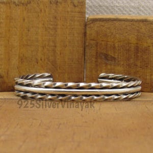 Puede incluir: Una pulsera de plata con un diseño de cuerda trenzada. La pulsera tiene una banda lisa y pulida en el centro y está estampada con el texto "925SilverVinayak". La pulsera se exhibe sobre una superficie de madera.