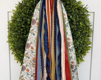 May include: A round green boxwood wreath adorned with a large decorative bow featuring a floral pattern in orange, blue, and green. Long, cascading ribbons in solid navy blue, mustard yellow, rust red, and light blue hang from the center.