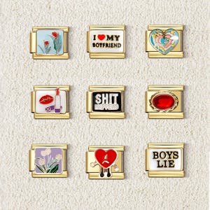 Op de afbeelding: Negen goudkleurige bedelarmbanden met diverse ontwerpen. De bedels omvatten een hart met "I MY BOYFRIEND", een hart met een verdrietig gezicht, een rode edelsteen, een lippenstift en lippen, en de woorden "BOYS LIE" en "SHIT HAPPENS".