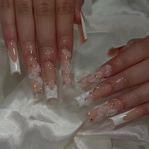 Könnte beinhalten: Lange, quadratische Acrylnägel mit French-Tip-Design. Die Nägel haben eine nudefarbene Basis mit weißen Spitzen, verziert mit 3D-Blütenakzenten in Weiß und goldenen Strasssteinen. Die Nägel sind lang und elegant.