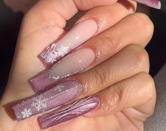 May include: Long, square-shaped artificial nails in shades of pink and purple. Some nails feature a French tip design, while others have silver glitter accents and white snowflake designs. The nails are a winter-themed manicure.
