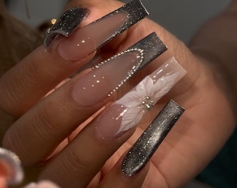 May include: Long, square-shaped acrylic nails. Some nails feature a glittery black design on the tips, while others have a clear base with white floral accents and rhinestones. The nails are a light pink color.
