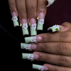 May include: Long, square-shaped artificial nails with a light green and pink color scheme. Each nail features a white flower and gold accents. The nails are decorated with 3D floral designs and small gold embellishments.